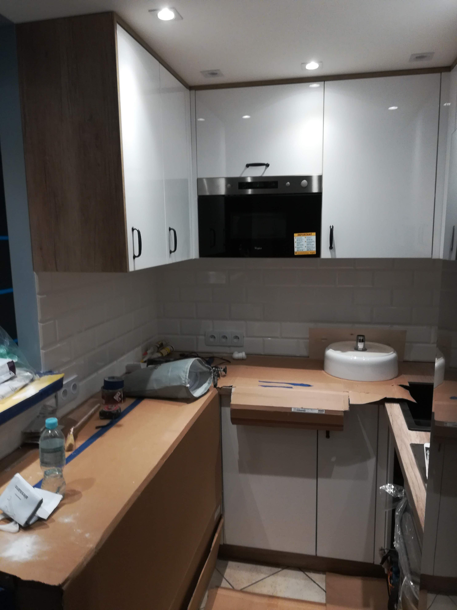 Montaż kuchni: białe szafki wiszące i stojące, zabudowa piekarnika, biała umywalka nablatowa, kartonowe zabezpieczenia blatu, widoczne narzędzia i akcesoria instalacyjne.