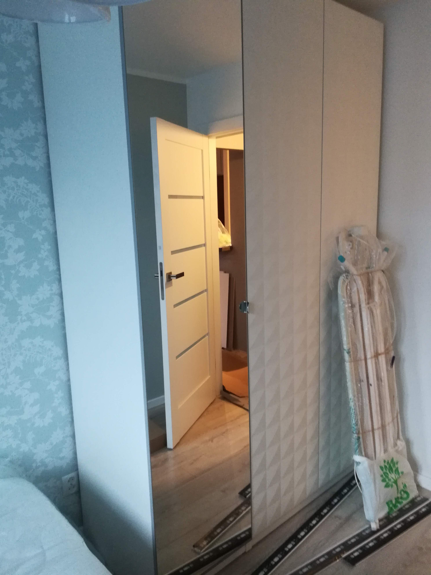 Biała szafa z lustrzanymi drzwiami, w których odbija się otwarte drzwi do innego pomieszczenia. Obok szafy leżą elementy montażowe w folii i torbie.