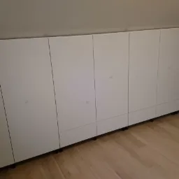 Biała szafa wnękowa z minimalistycznymi uchwytami na poddaszu, widok z dołu na drewnianą podłogę i fragment niebieskiej ściany.