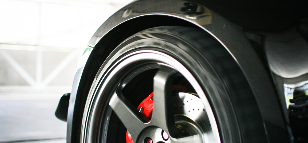 Błyszczący, czarny błotnik samochodu z widocznym szarym felgią aluminiowym i czerwonym zaciskiem hamulcowym, na tle jasnego garażu.