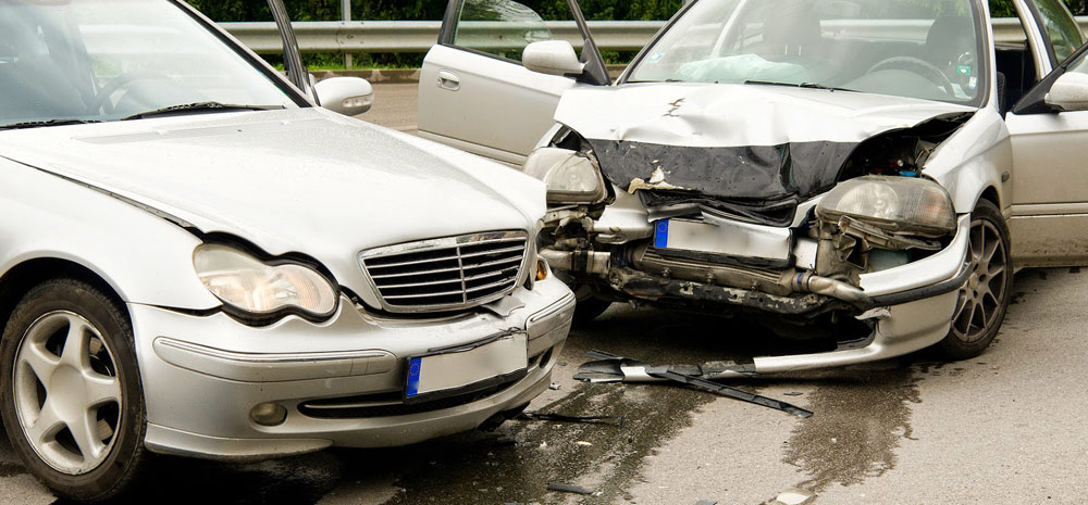 Dwa srebrne samochody po kolizji czołowej, widoczne uszkodzenia karoserii i elementy rozbite na jezdni.