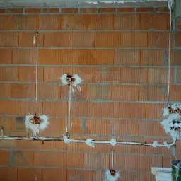 Wewnętrzna ściana z czerwonej cegły z widocznymi przewodami elektrycznymi wychodzącymi z puszek instalacyjnych, częściowo zamurowanych gipsem, widok na okno z ramą Salamander i puszkę farby.