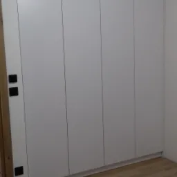 Biała szafa wnękowa z gładkimi frontami, zamontowana od podłogi do sufitu, z widocznymi włącznikami światła na ścianie obok i fragmentem odkurzacza na podłodze.