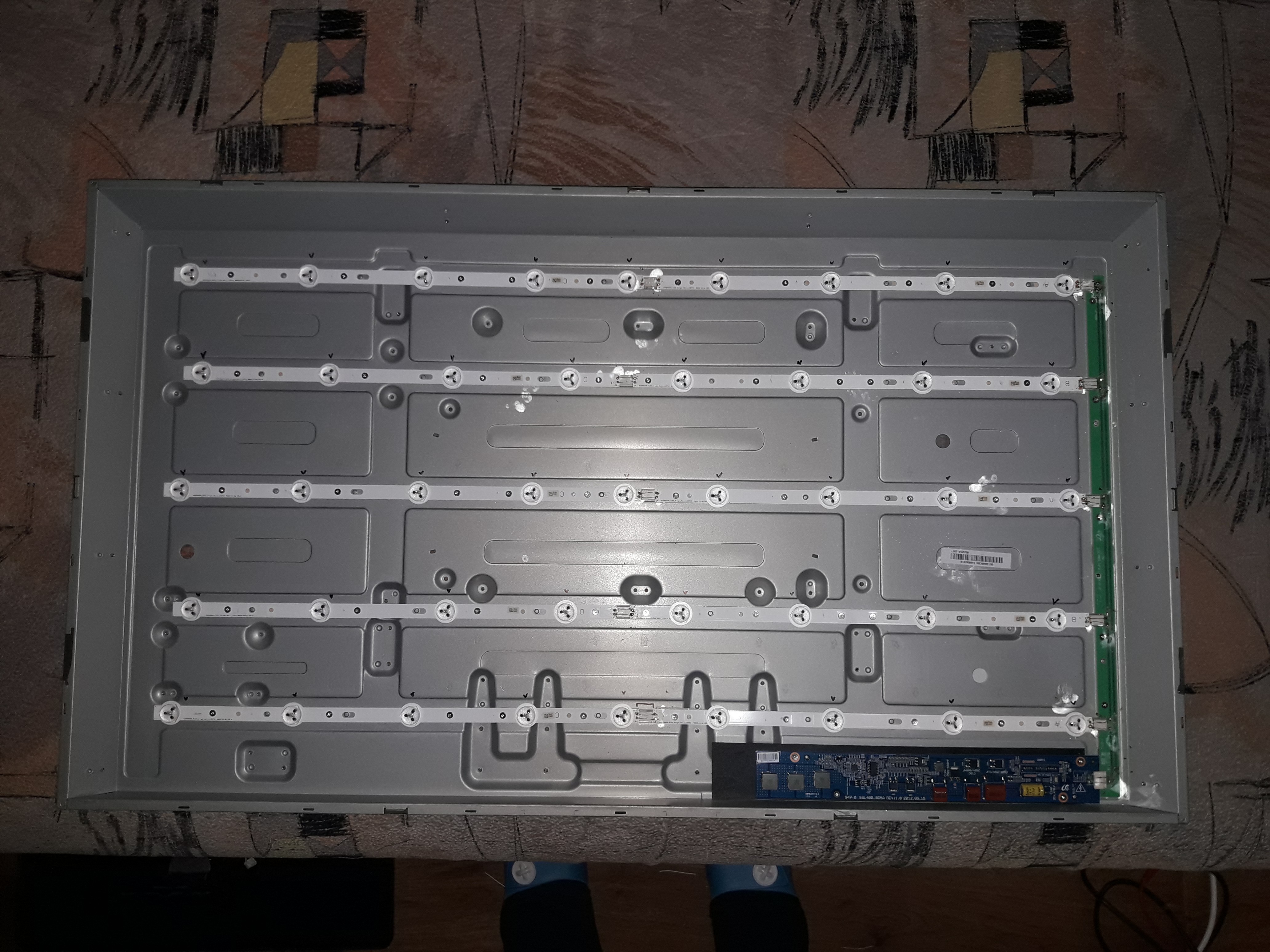 Wnętrze obudowy telewizora z widocznymi listwami LED podświetlenia matrycy oraz płytą główną, leżące na wzorzystej powierzchni.