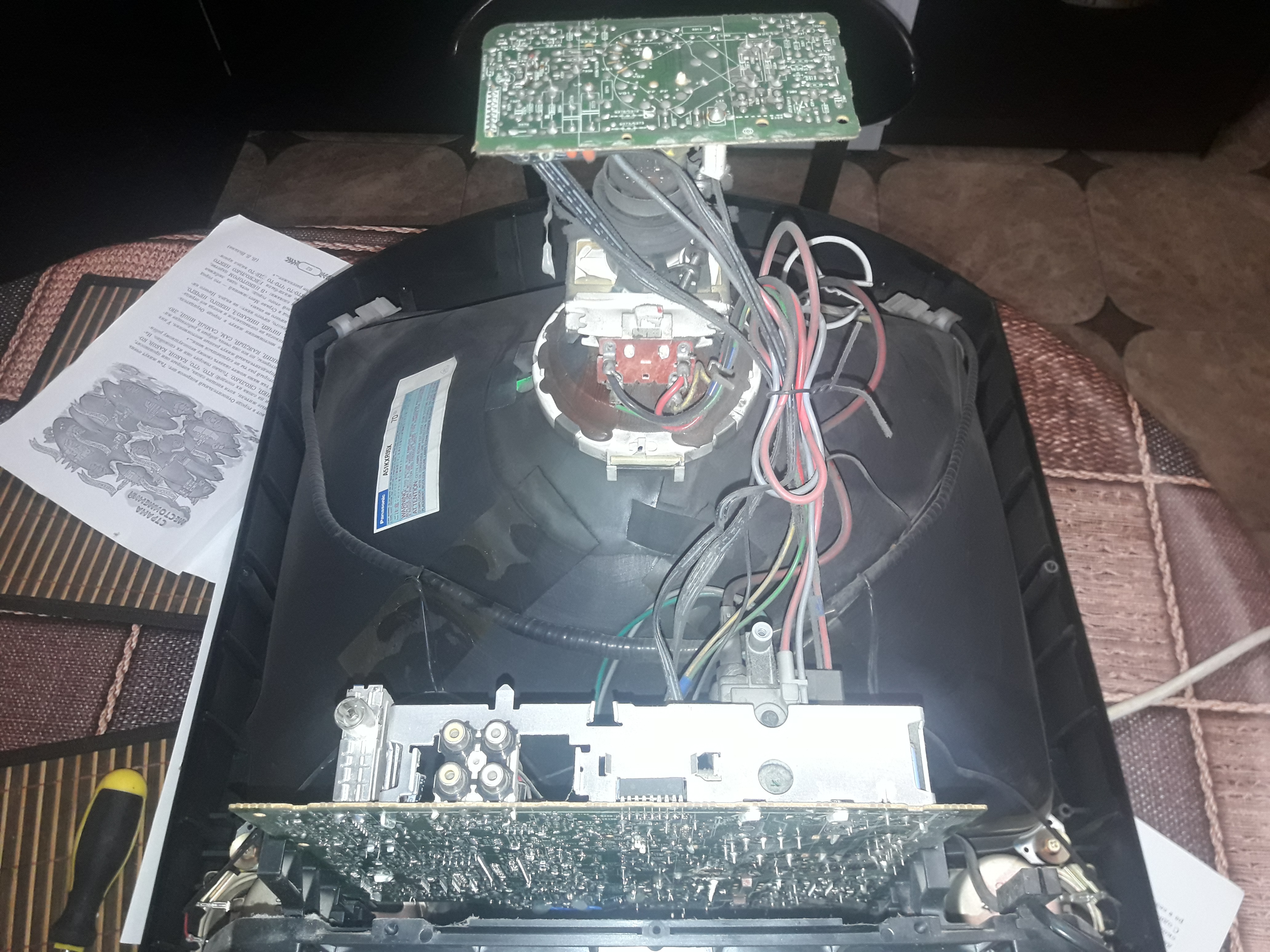 Wnętrze rozmontowanego telewizora kineskopowego z widocznymi płytkami drukowanymi, kablami i elementami elektroniki, obok leży kartka z tekstem w języku rosyjskim.