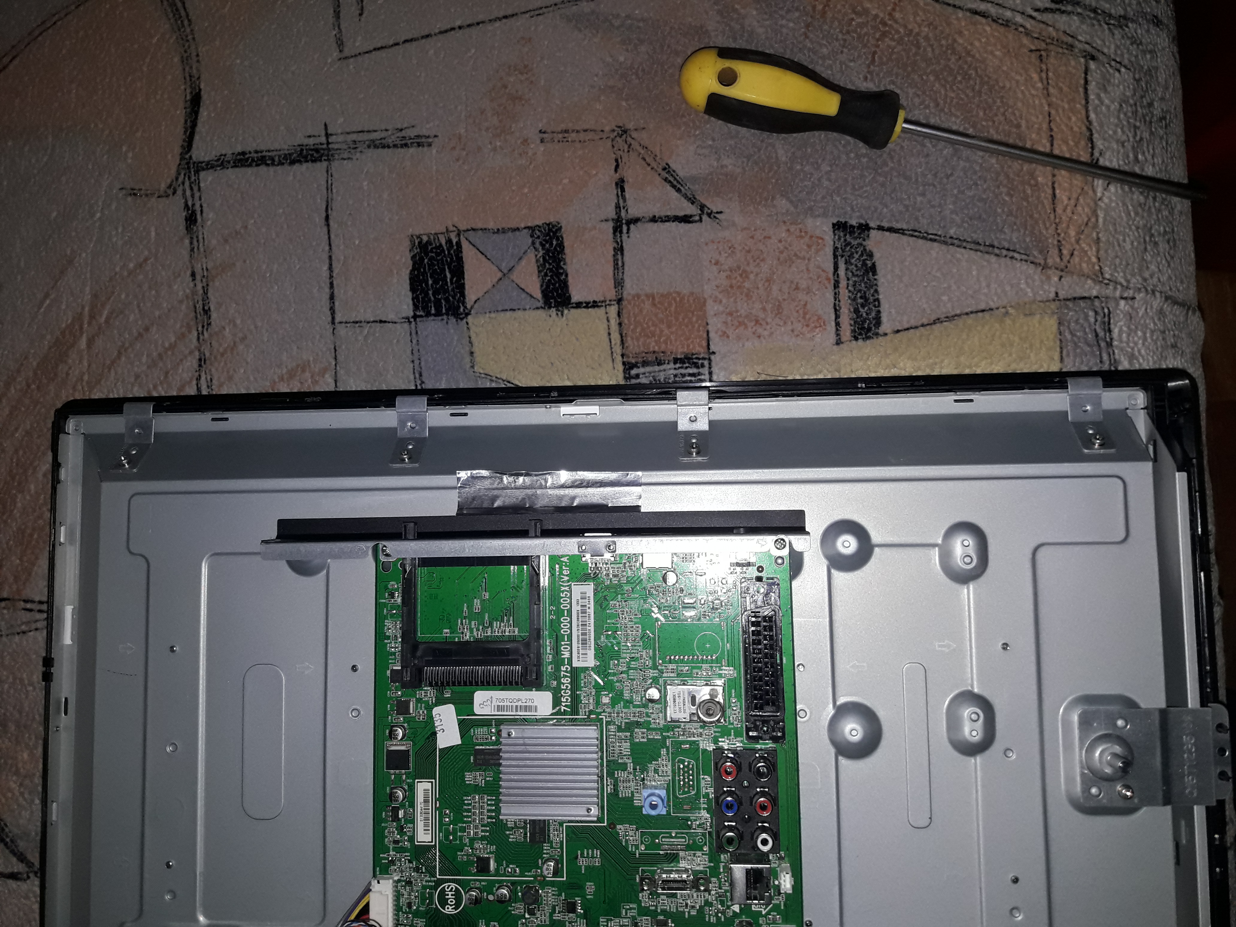 Otwarta obudowa telewizora z widoczną płytą główną i śrubokrętem z żółtą rączką leżącym obok, na tle wzorzystej tkaniny.