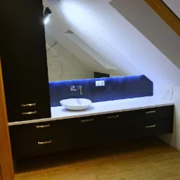 Nowoczesna łazienka na poddaszu ze skośnym sufitem, czarne szafki z białym blatem, umywalka nablatowa i podświetlenie LED za lustrem.