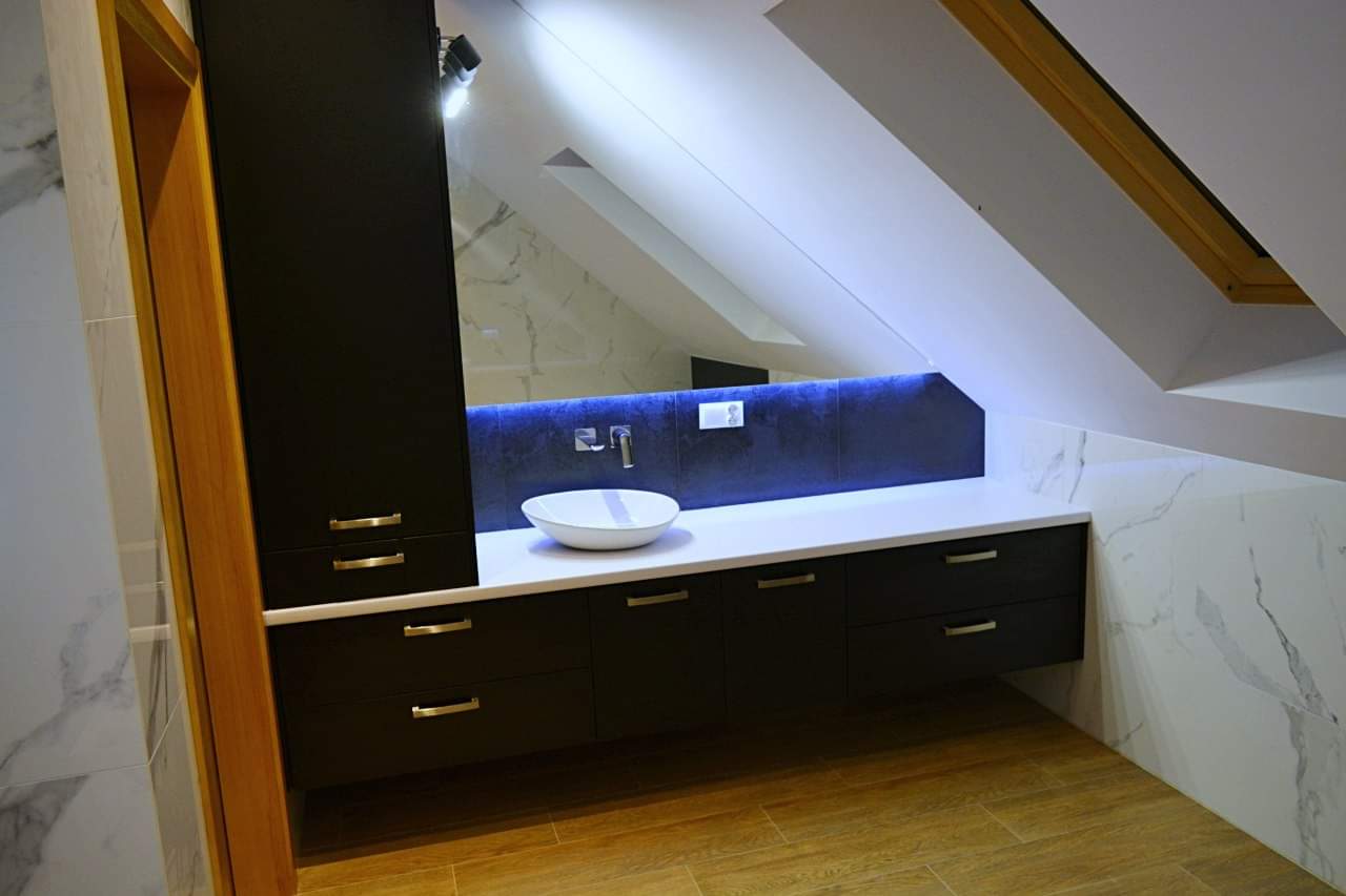 Nowoczesna łazienka na poddaszu ze skośnym sufitem, czarne szafki z białym blatem, umywalka nablatowa i podświetlenie LED za lustrem.