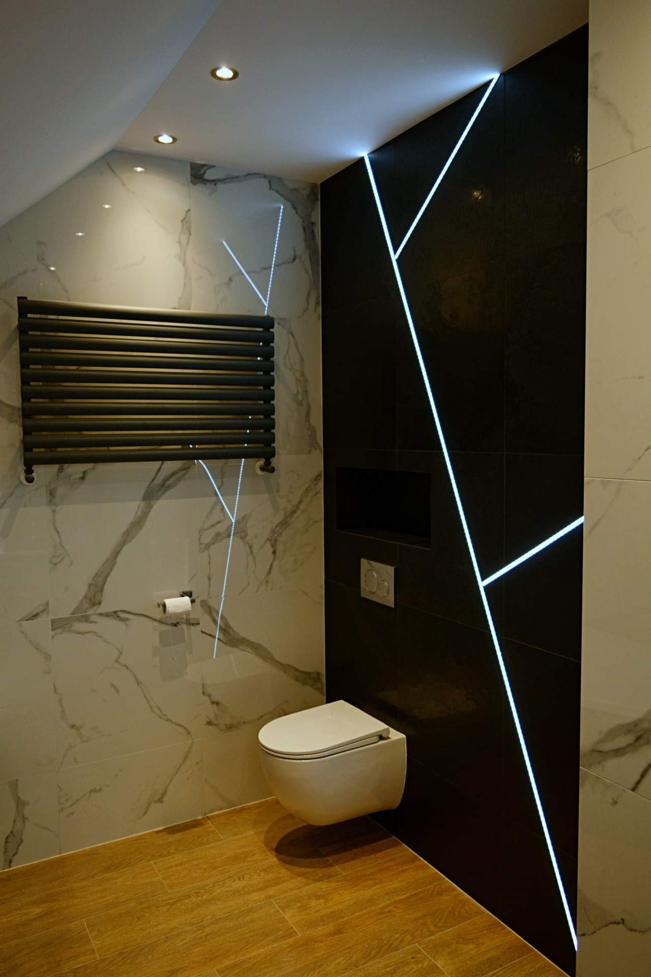 Nowoczesna łazienka z czarnymi i marmurowymi płytkami, podświetleniem LED w geometryczne wzory, wiszącą toaletą i grzejnikiem drabinkowym.