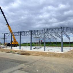 JOT Zakład Produkcyjny - Montaż stalowej konstrukcji hali z użyciem dźwigu na betonowej płycie fundamentowej, widok z zewnątrz w pochmurny dzień.