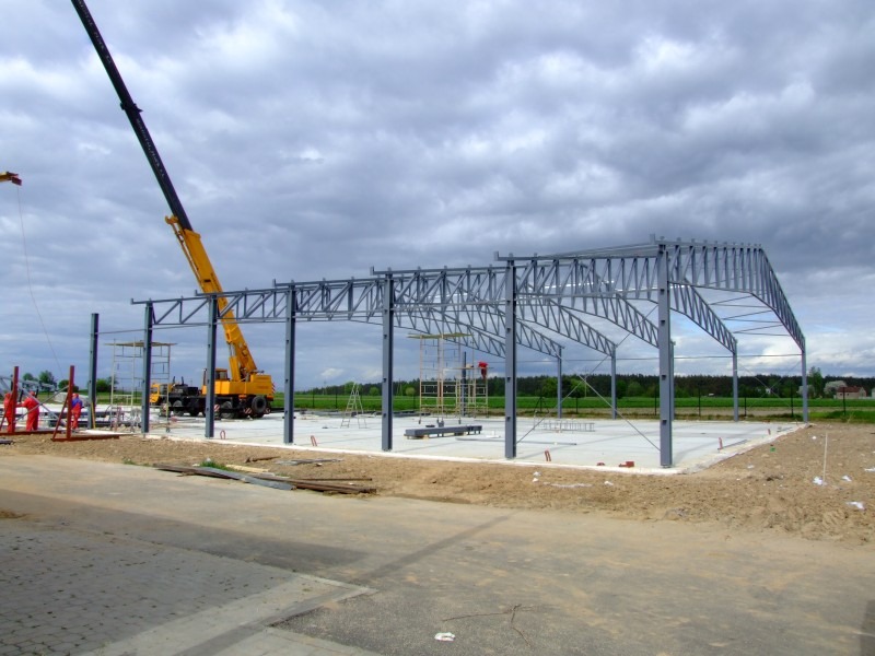 Montaż stalowej konstrukcji hali z użyciem dźwigu na betonowej płycie fundamentowej, widok z zewnątrz w pochmurny dzień.