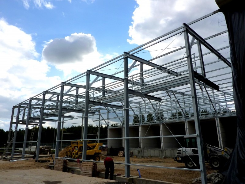 Stalowa konstrukcja hali w trakcie montażu, widoczne elementy nośne, słupy i belki, na tle błękitnego nieba z chmurami, z żółtą koparką i białym miniładowarką na placu budowy.