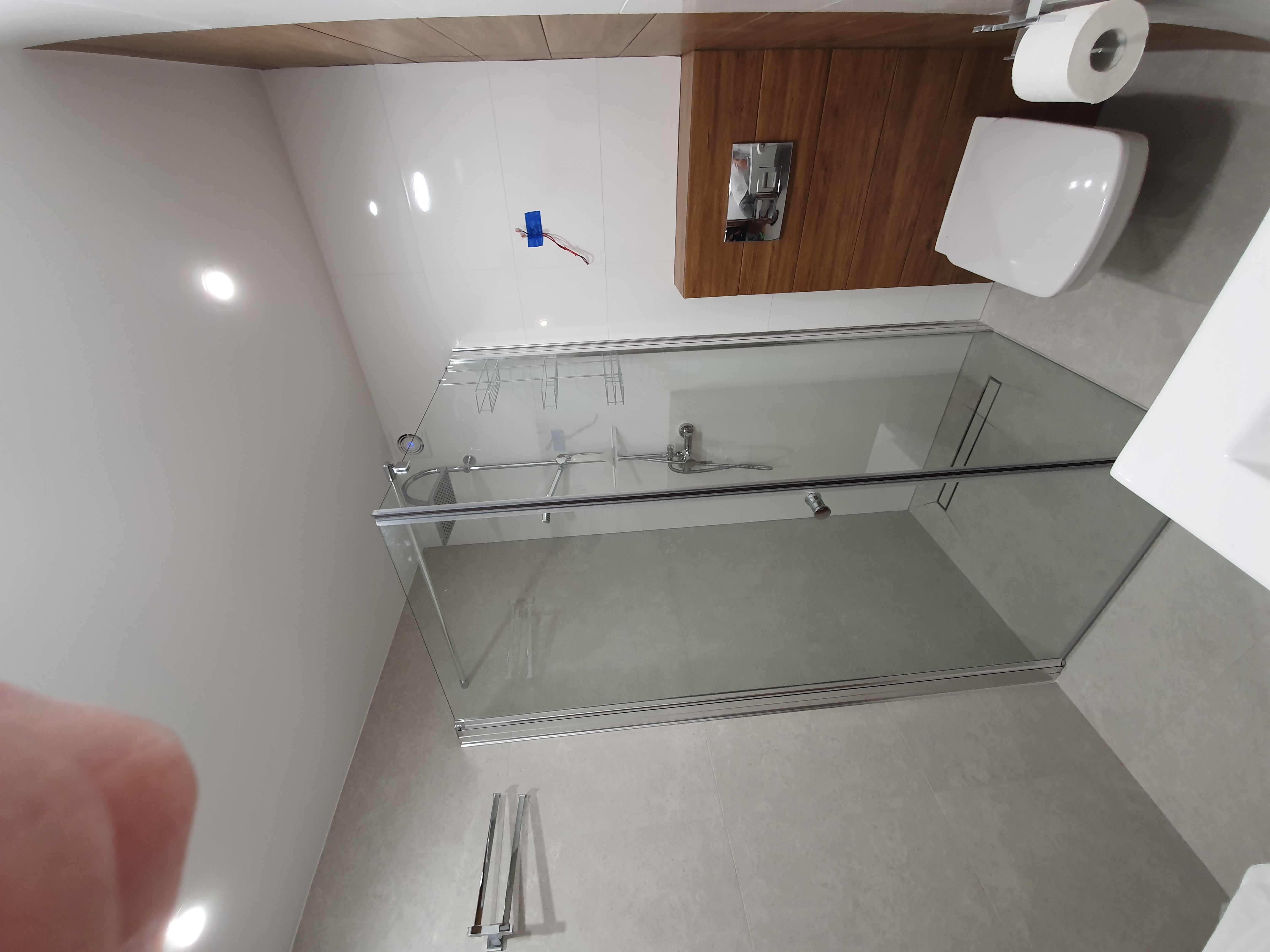 Nowoczesna kabina prysznicowa z przesuwnymi drzwiami, zamontowana w wykończonej łazience z białymi płytkami i elementami drewnopodobnymi, widoczny także podwieszany sedes i umywalka.