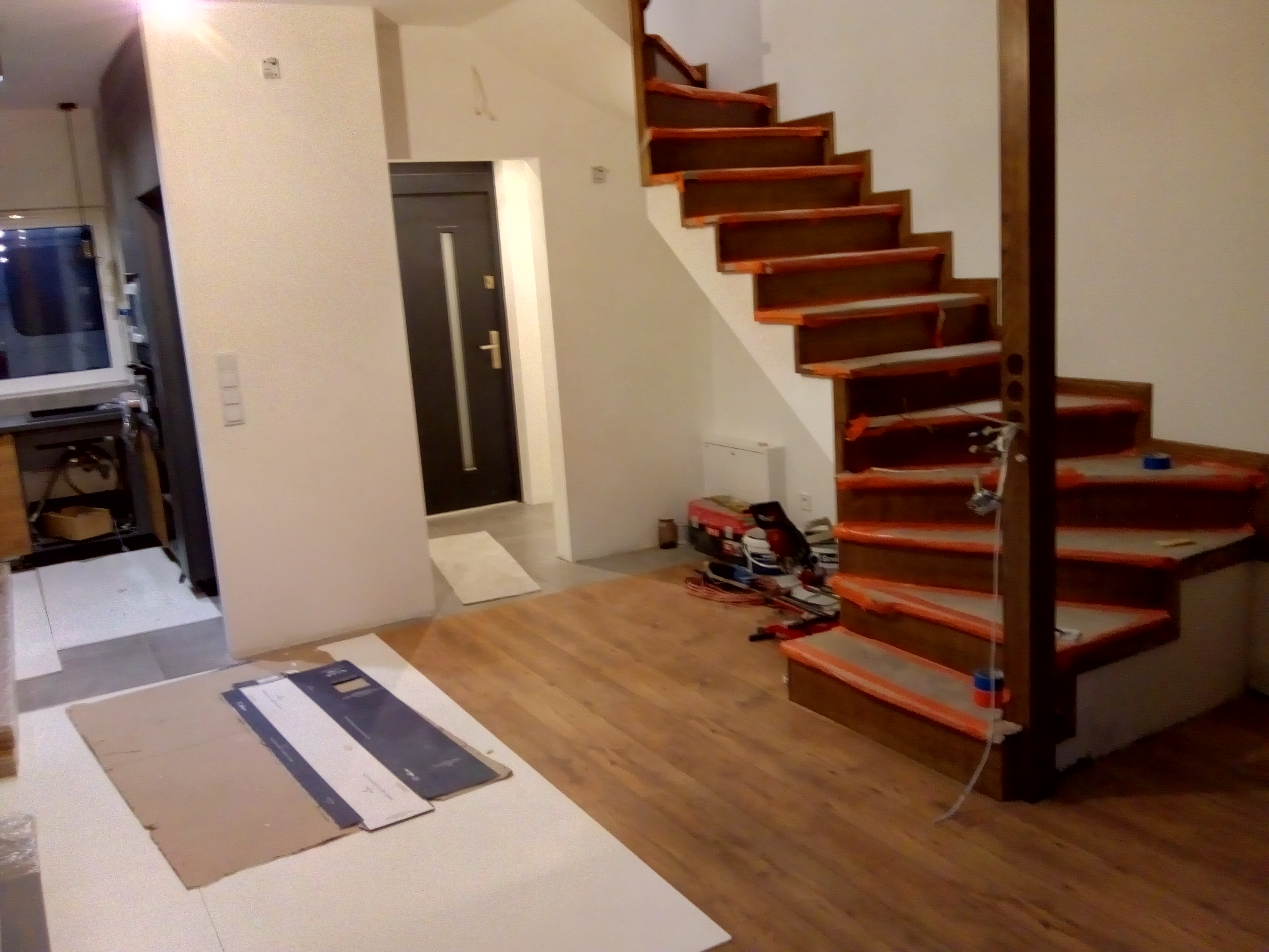 Wnętrze w trakcie wykańczania: drewniane schody zabezpieczone taśmą malarską, panele podłogowe, drzwi wewnętrzne i narzędzia w tle.