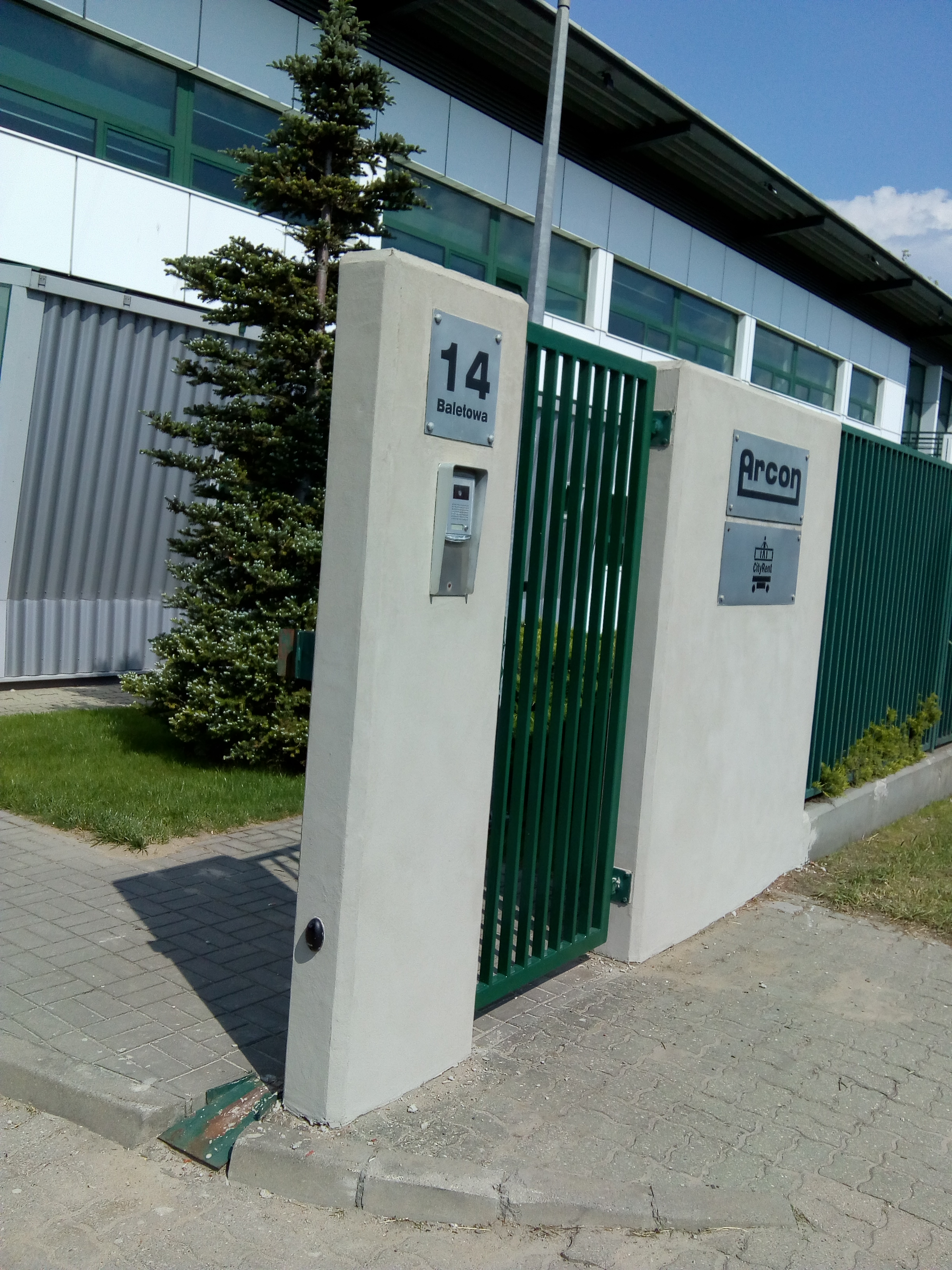 Wejście do biura z zieloną bramą, numerem 14 i logo firmy Arcon na betonowym słupku, obok rośnie mała choinka.