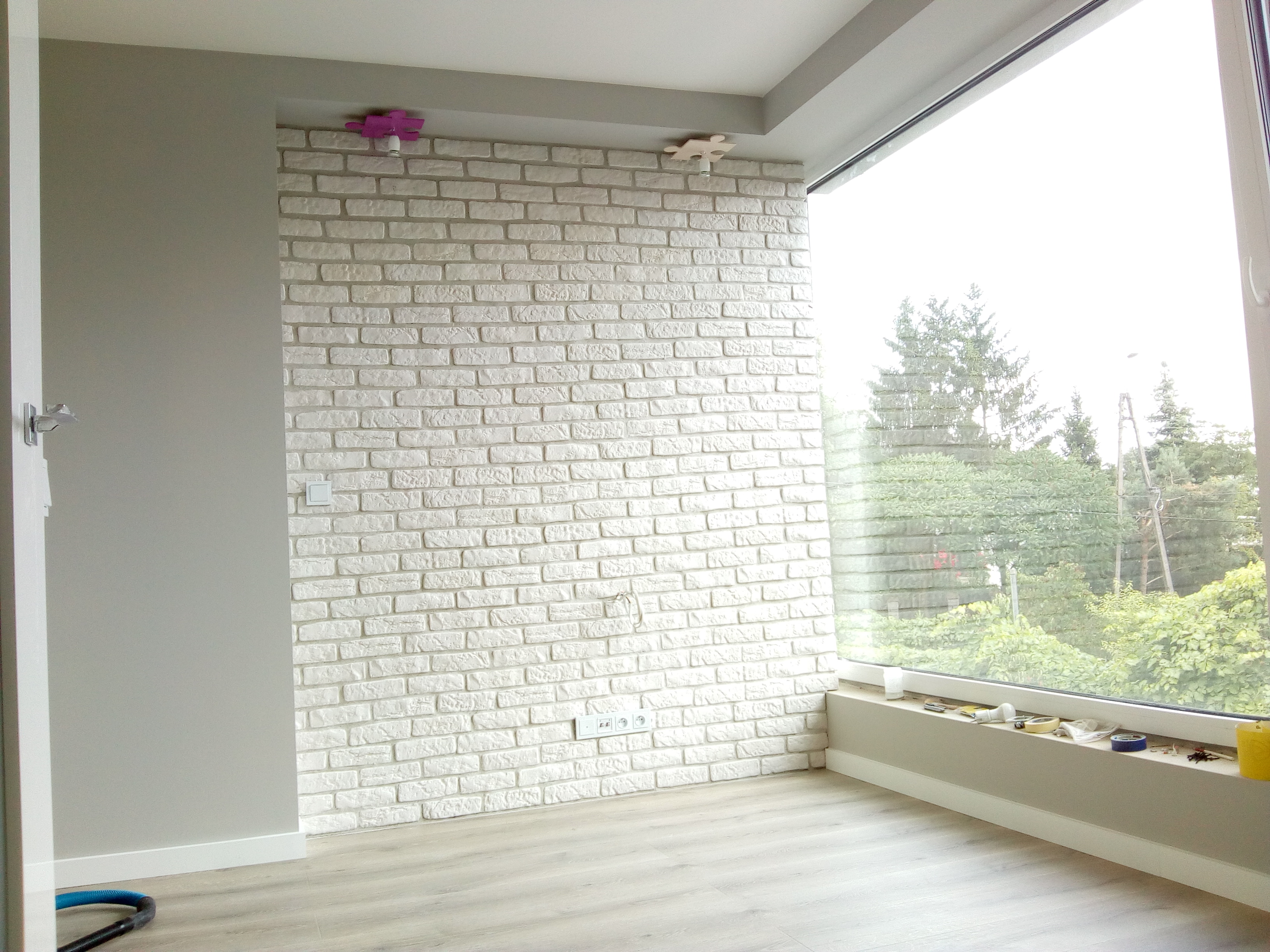 Świeżo wykończone wnętrze z białą cegłą na ścianie, widoczne gniazdka elektryczne, okno z widokiem na zieleń i lampy sufitowe w kształcie puzzli.