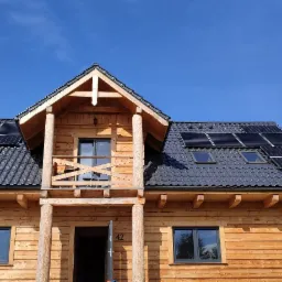 Drewniany dom z ciemną dachówką i panelami słonecznymi zamontowanymi na dachu, widok z zewnątrz w słoneczny dzień.