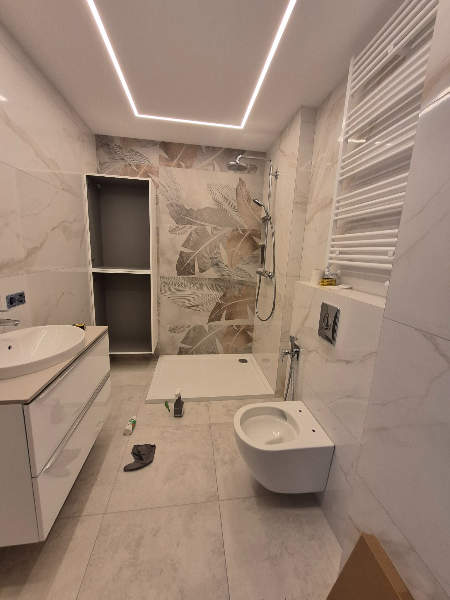 Nowoczesna łazienka w trakcie wykańczania: białe płytki imitujące marmur, podwieszana toaleta, kabina prysznicowa typu walk-in, grzejnik drabinkowy i oświetlenie LED.