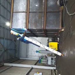 Spawacz w kombinezonie i masce spawalniczej, stojący na aluminiowej drabinie Krause, spawa metalową konstrukcję w hali przemysłowej, widoczne iskry i żółty spawarka ESAB.