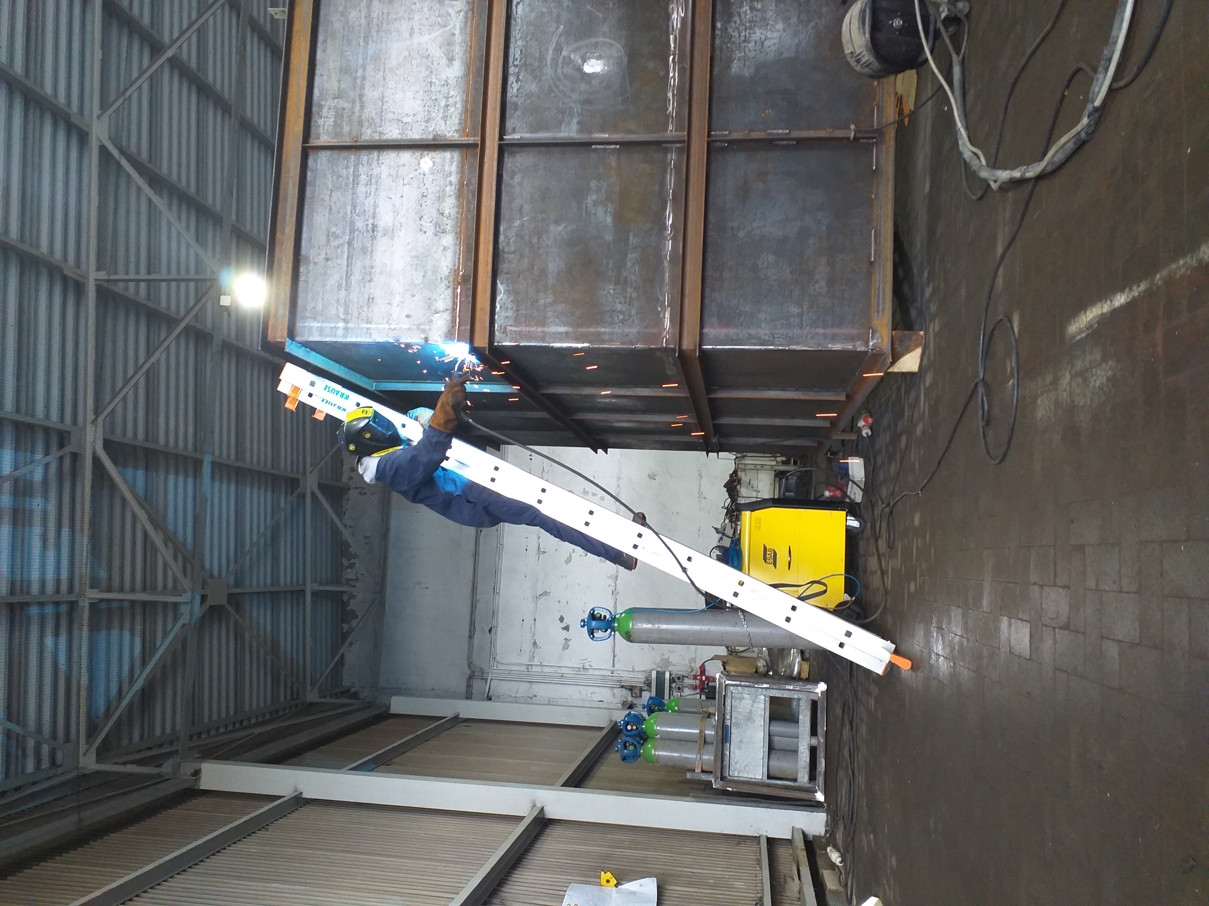 Spawacz w kombinezonie i masce spawalniczej, stojący na aluminiowej drabinie Krause, spawa metalową konstrukcję w hali przemysłowej, widoczne iskry i żółty spawarka ESAB.