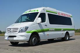 Biały bus Mercedes Sprinter z zielonymi akcentami i logo firmy TRANS-A na tle błękitnego nieba i betonowej nawierzchni.