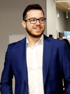 Portret uśmiechniętego mężczyzny w okularach i niebieskiej marynarce, stojącego na jasnym tle z elementami wystawienniczymi w tle.