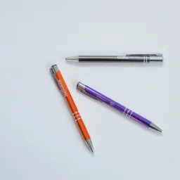 Trzy metalowe długopisy z nadrukowanym logo firmowym w kolorach pomarańczowym, fioletowym i srebrnym, leżące na białym tle.