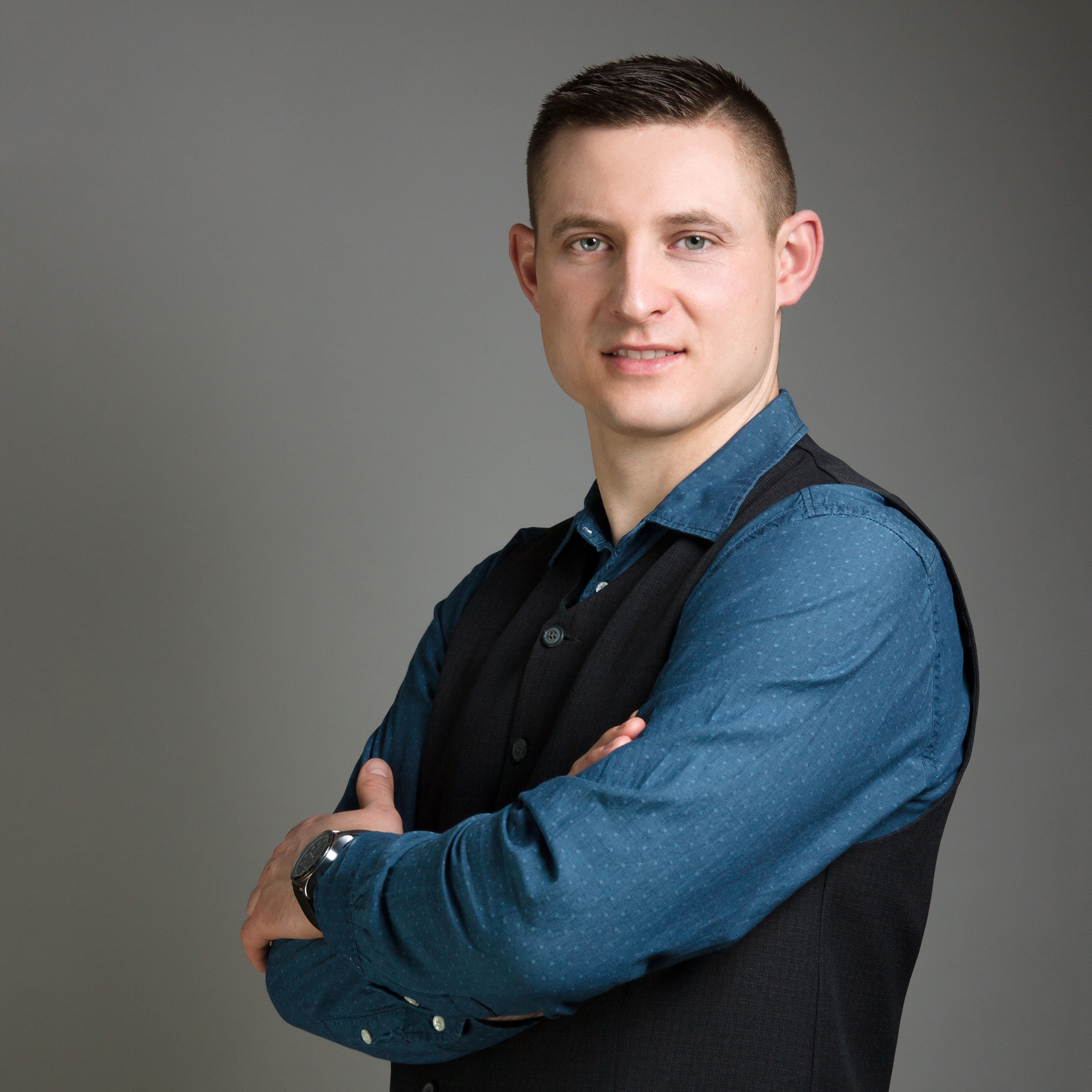 Portret mężczyzny w niebieskiej koszuli i czarnej kamizelce, z zegarkiem na lewym nadgarstku, stojącego na szarym tle z założonymi rękami.