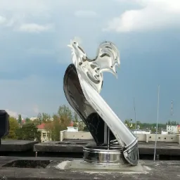 Błyszcząca, metalowa rzeźba koguta na kominie, z widocznym tłem miejskim i pochmurnym niebem.