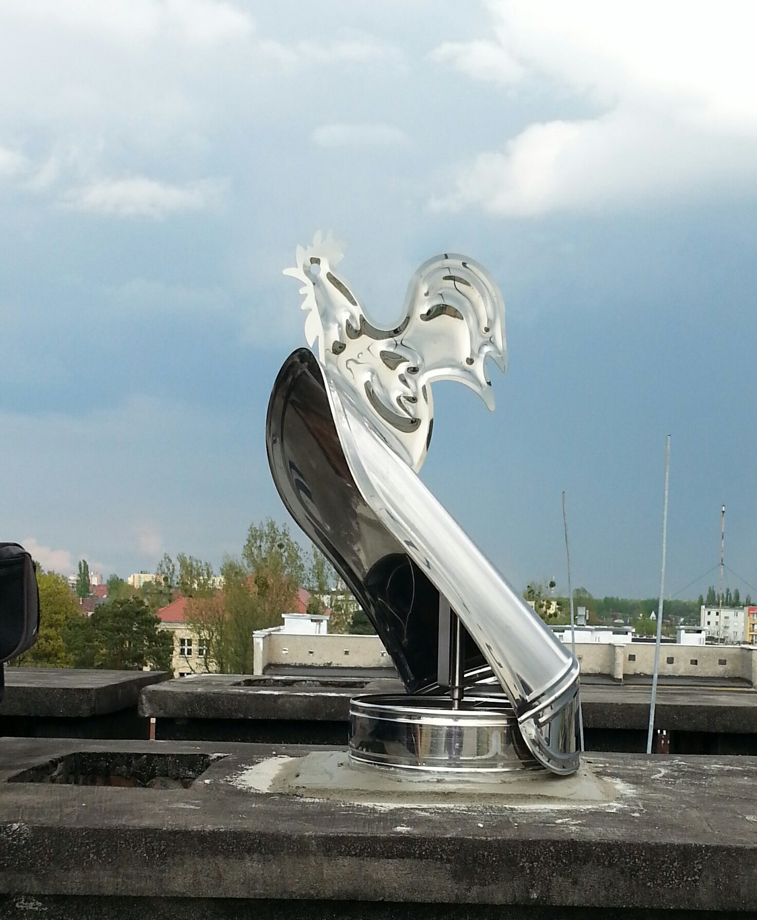 Błyszcząca, metalowa rzeźba koguta na kominie, z widocznym tłem miejskim i pochmurnym niebem.