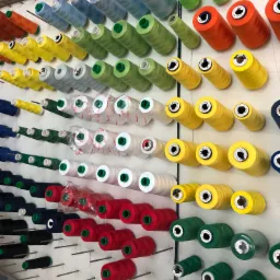 Rzędy kolorowych nici na szpulach, umieszczonych na metalowych prętach przy ścianie, widoczne etykiety producentów i oznaczenia grubości nici.