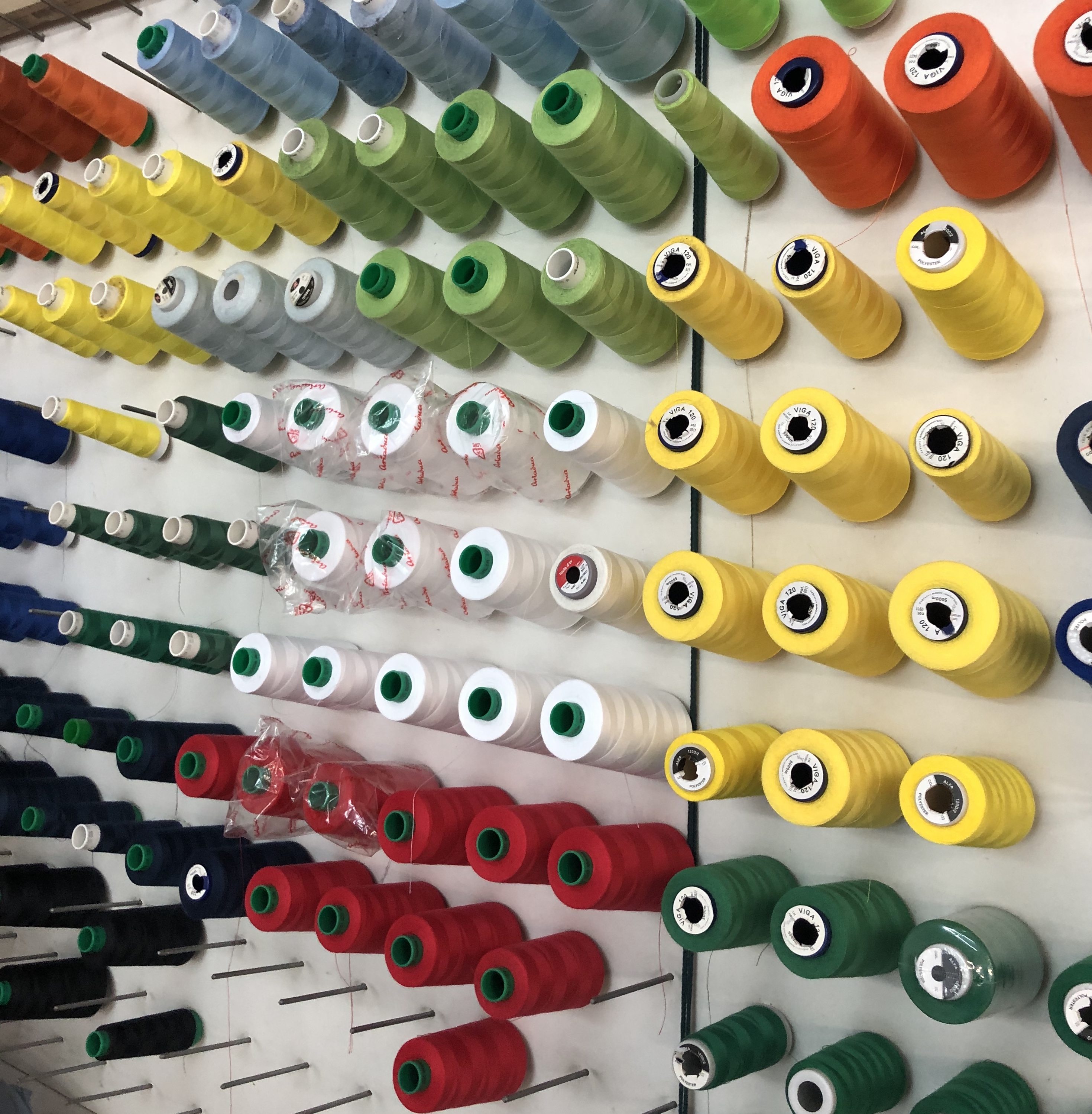 Rzędy kolorowych nici na szpulach, umieszczonych na metalowych prętach przy ścianie, widoczne etykiety producentów i oznaczenia grubości nici.