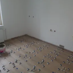 Układanie drewnopodobnych paneli z użyciem klipsów dystansowych, widoczne puste puszki po materiałach budowlanych i otwory w ścianie.