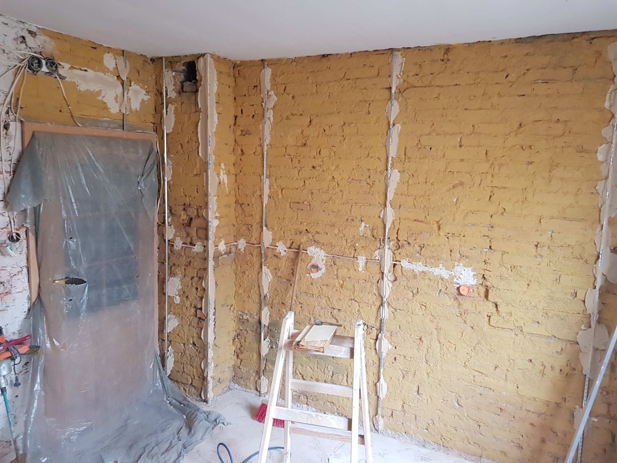 Przygotowanie ścian do nałożenia gładzi: surowe ściany z cegły pokryte warstwą zaprawy, metalowe profile pionowe przytwierdzone do ścian, drabina z deską i narzędziami w pomieszczeniu...