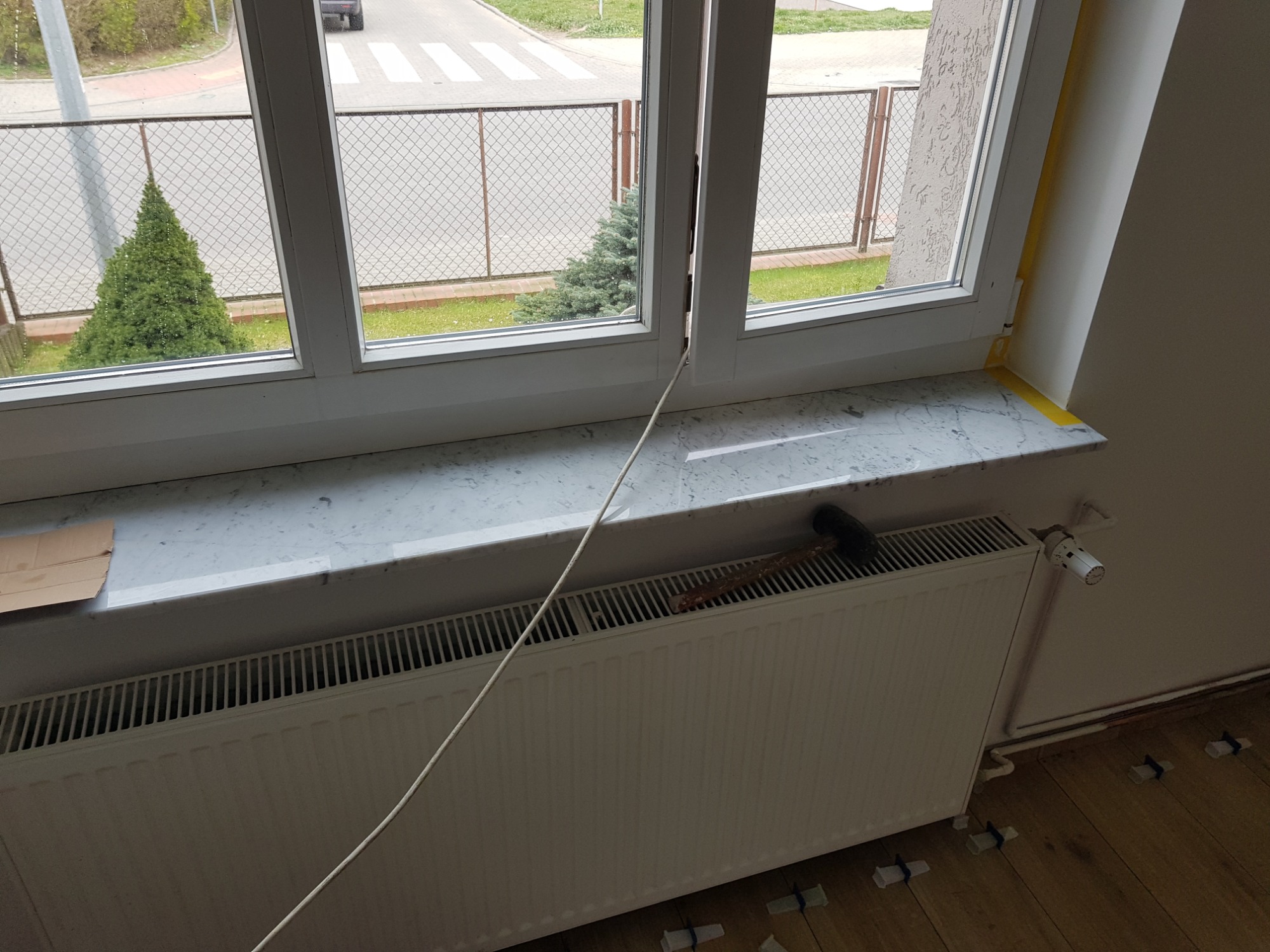 Nowy marmurowy parapet pod białym oknem z widokiem na ulicę, z grzejnikiem pod spodem, młotek na grzejniku i zabezpieczone taśmą panele podłogowe.