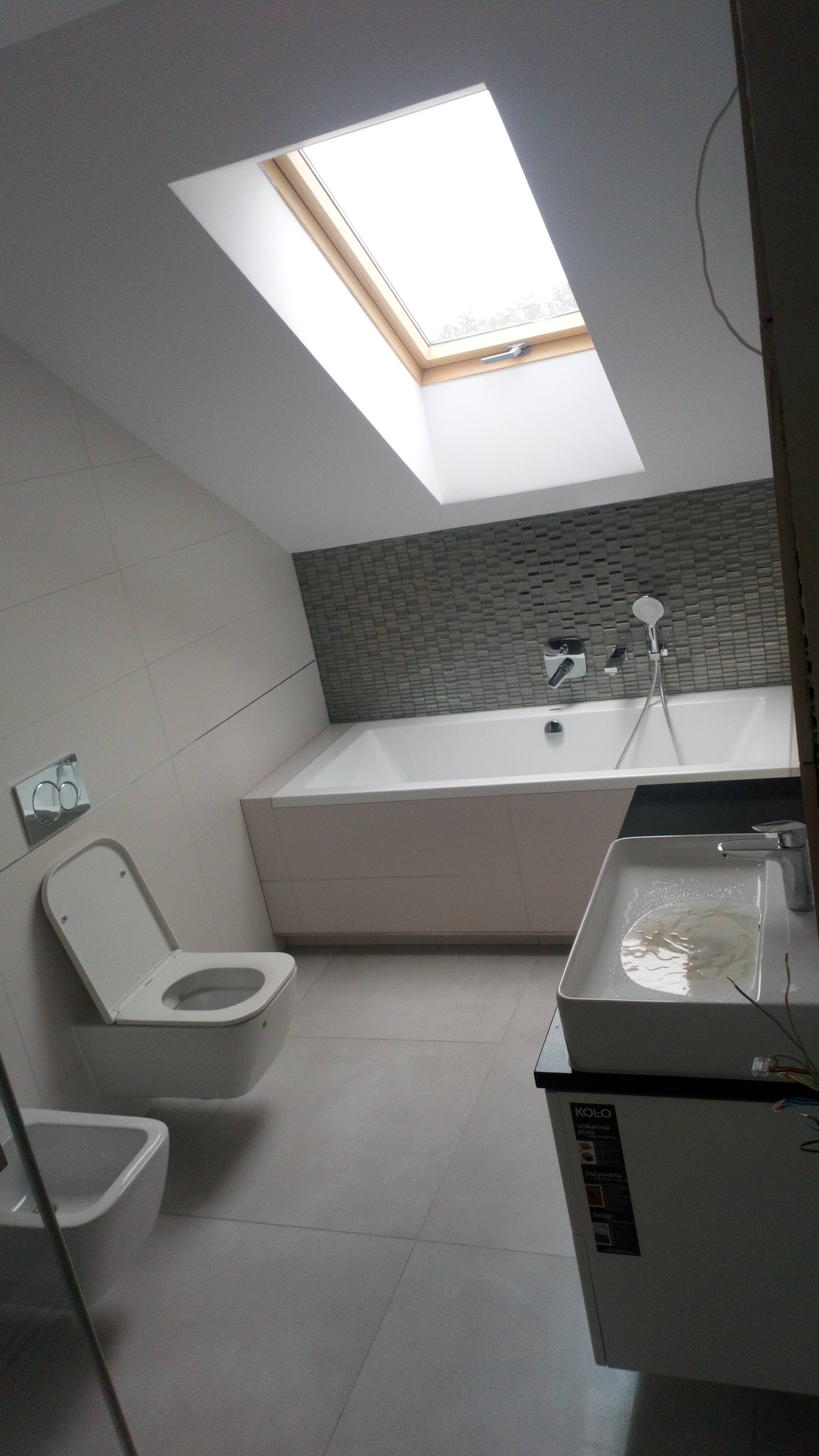 Nowoczesna łazienka na poddaszu ze skośnym sufitem i oknem dachowym, biała wanna obudowana płytami, umywalka nablatowa z czarnym blatem, wisząca toaleta z chromowanym przyciskiem spłukiwania, szare...