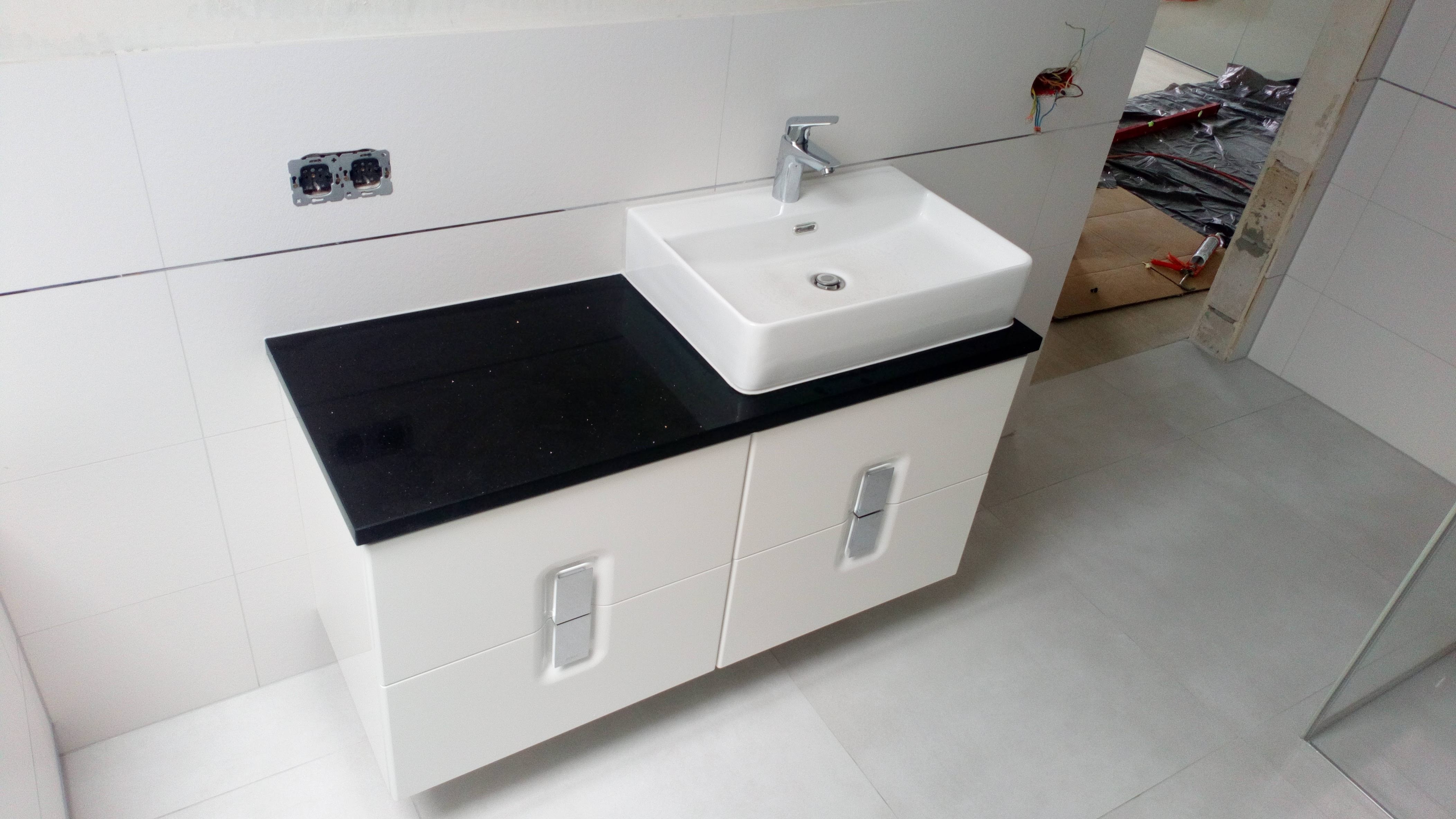 Biała, prostokątna umywalka nablatowa z chromowaną baterią na czarnym blacie szafki łazienkowej z dwiema szufladami, widoczne niedokończone gniazdka elektryczne i zabezpieczona folią podłoga w tle.