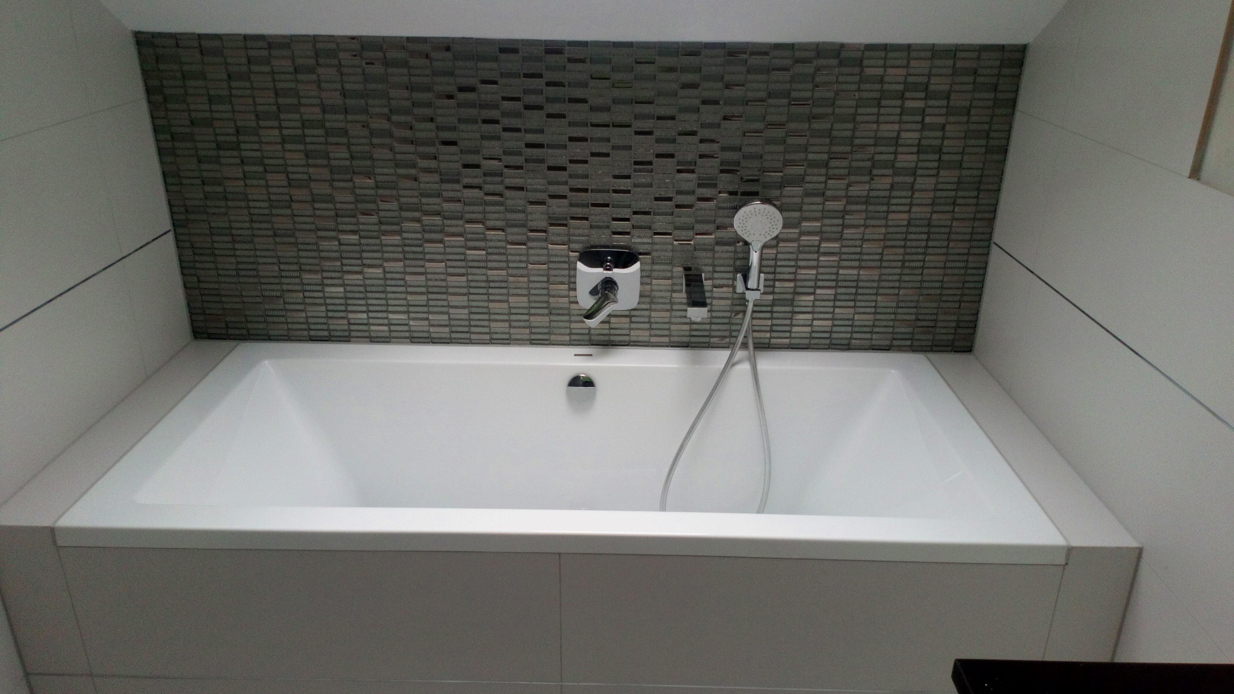 Biała wanna obudowana płytkami, na ścianie mozaika z szarych płytek, bateria wannowa z prysznicem.