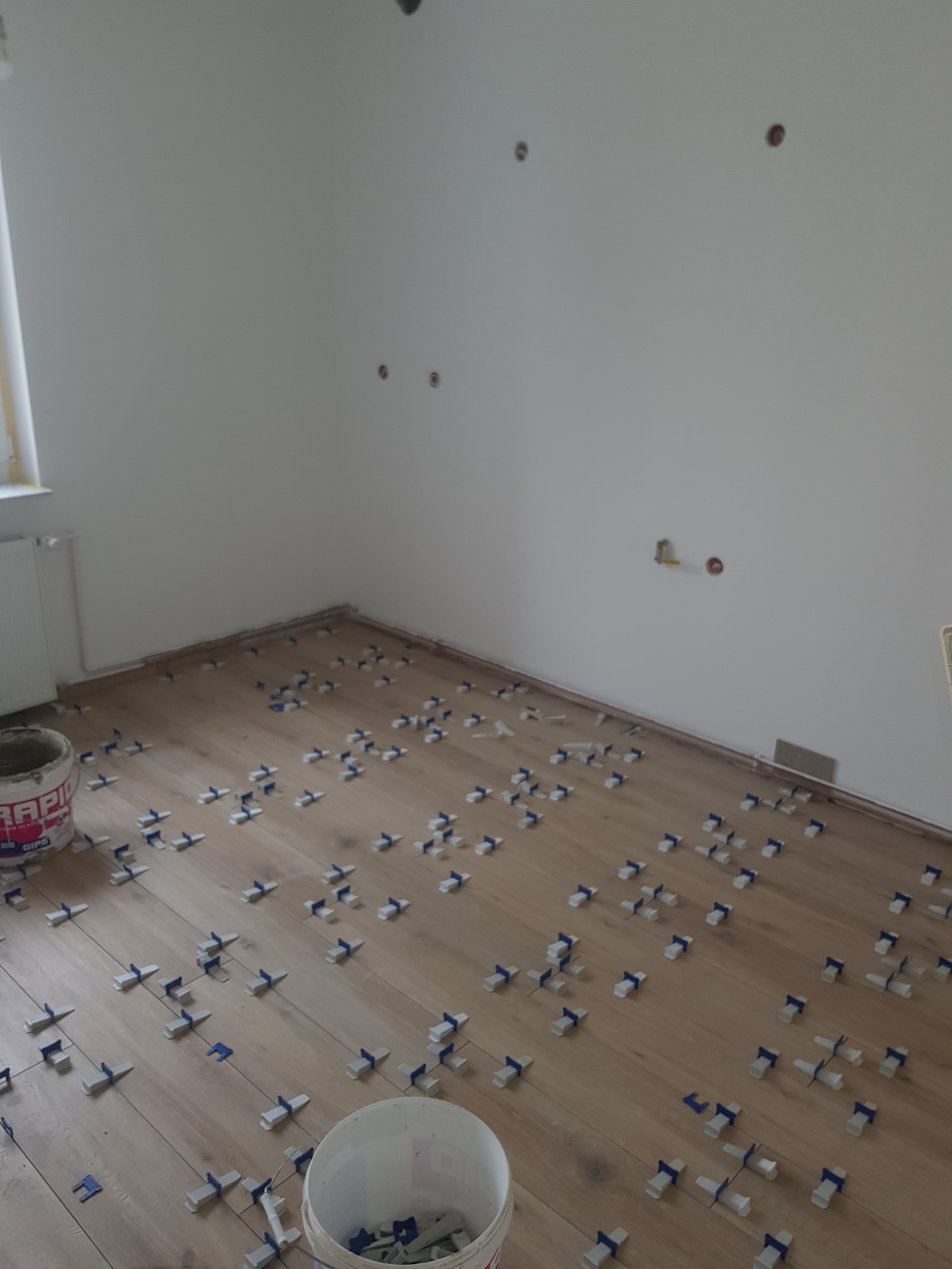 Układanie drewnopodobnych paneli z użyciem klipsów dystansowych, widoczne puste puszki po materiałach budowlanych i otwory w ścianie.