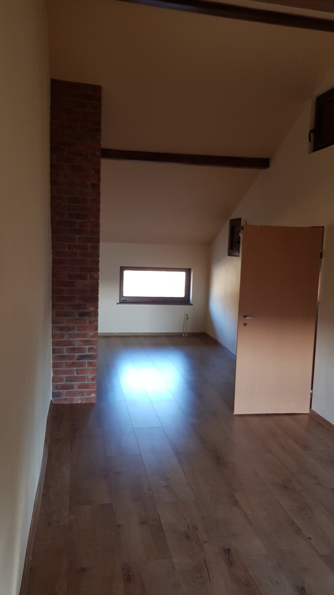 Pokój na poddaszu po remoncie w Koszalinie, z nową podłogą z paneli, ceglanym filarem i widocznym oknem dachowym.