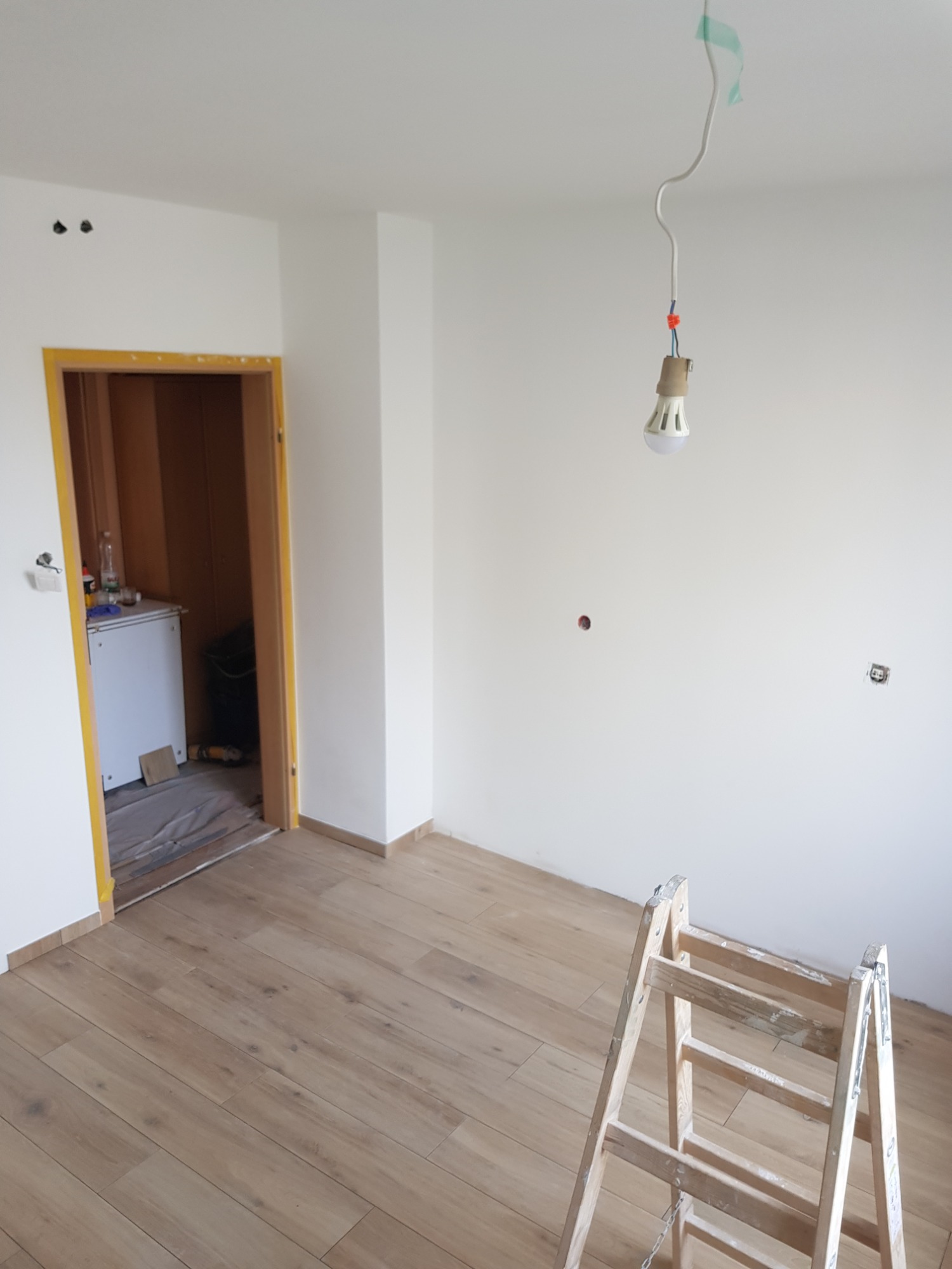 Pokój w trakcie prac wykończeniowych: świeżo pomalowane białe ściany, drewniana podłoga, drabina malarska i tymczasowe oświetlenie z wiszącą żarówką.