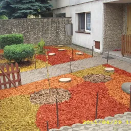 Ogród przydomowy z rabatami wysypanymi korą w różnych kolorach, niskie krzewy, małe drzewko podwiązane do palika, pocięte pnie drzew jako dekoracja.