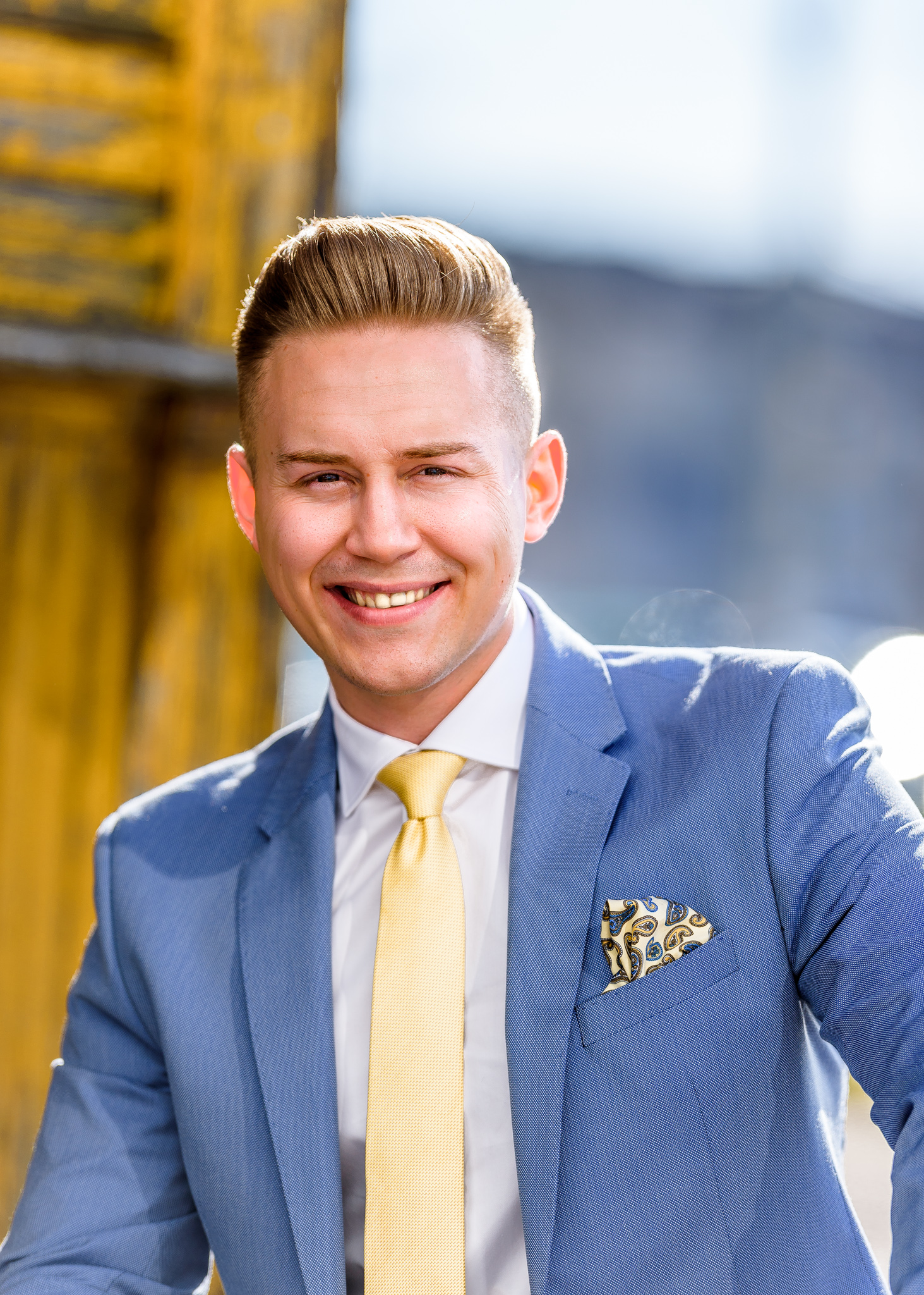 Uśmiechnięty młody mężczyzna w błękitnym garniturze z żółtym krawatem i wzorzystą poszetką, pozujący na tle rozmytej architektury miejskiej.