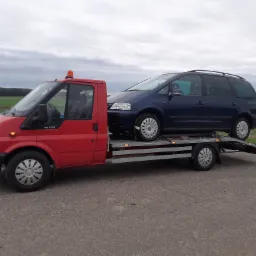 Czerwona laweta z niebieskim vanem Volkswagen Sharan na platformie, zabezpieczonym pasami transportowymi, na tle pochmurnego nieba i zielonych pól.