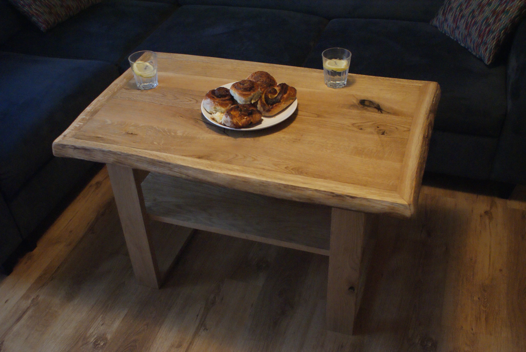 Stolik kawowy z blatem z surowego drewna, na którym leżą ciastka i stoją dwie szklanki z wodą i cytryną, na tle ciemnej kanapy.
