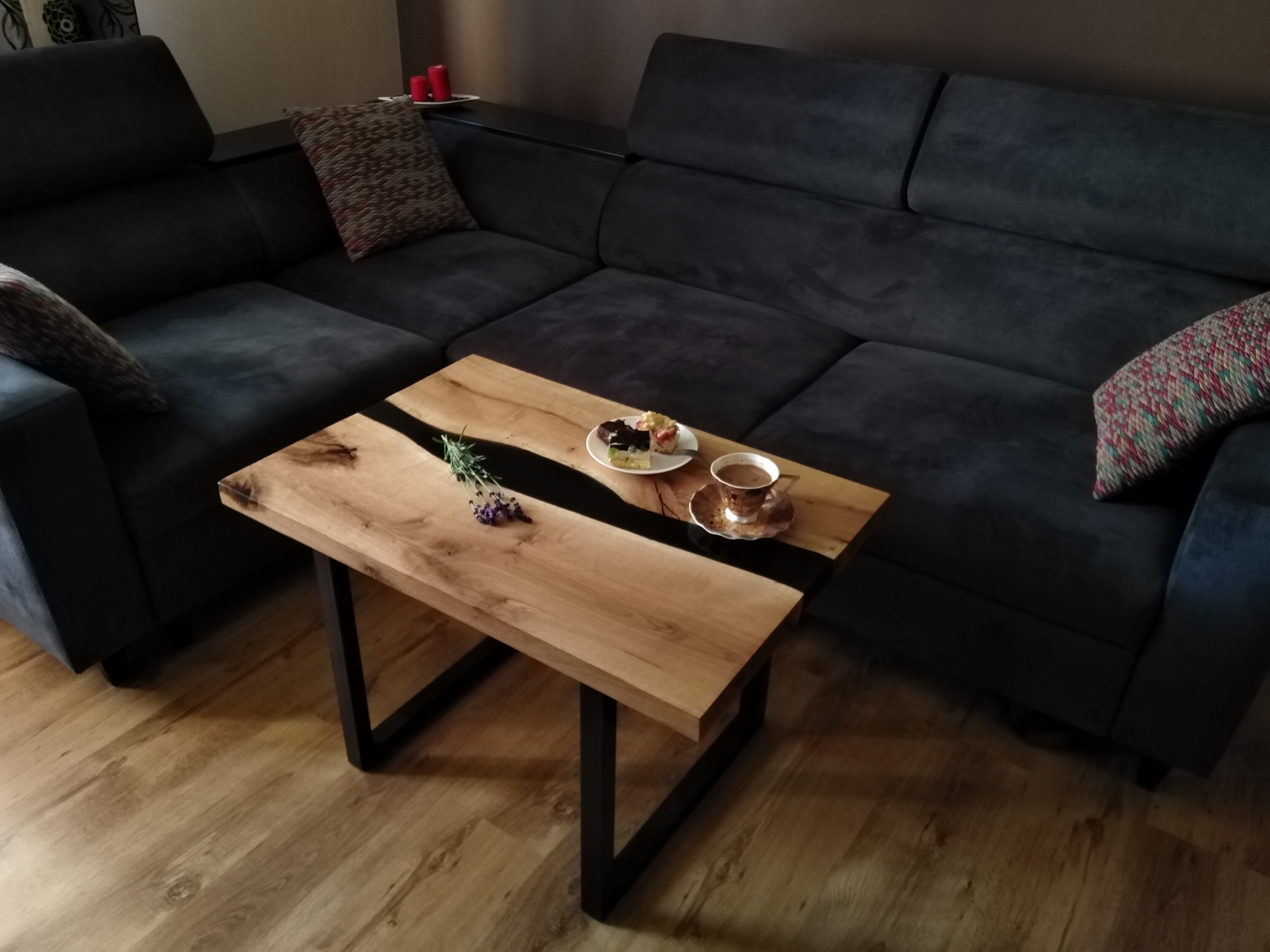 Stolik kawowy z blatem z jasnego drewna z żywicą epoksydową w kolorze czarnym, na którym leżą ciastka, filiżanka kawy i gałązka lawendy, na tle szarej, narożnej kanapy.