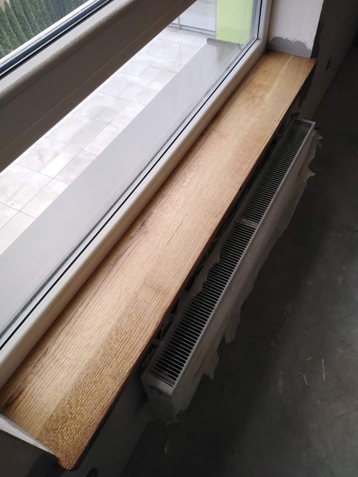 Zamontowany drewniany parapet o jasnym odcieniu drewna, widoczny grzejnik pod parapetem, surowy stan wykończenia wnętrza.