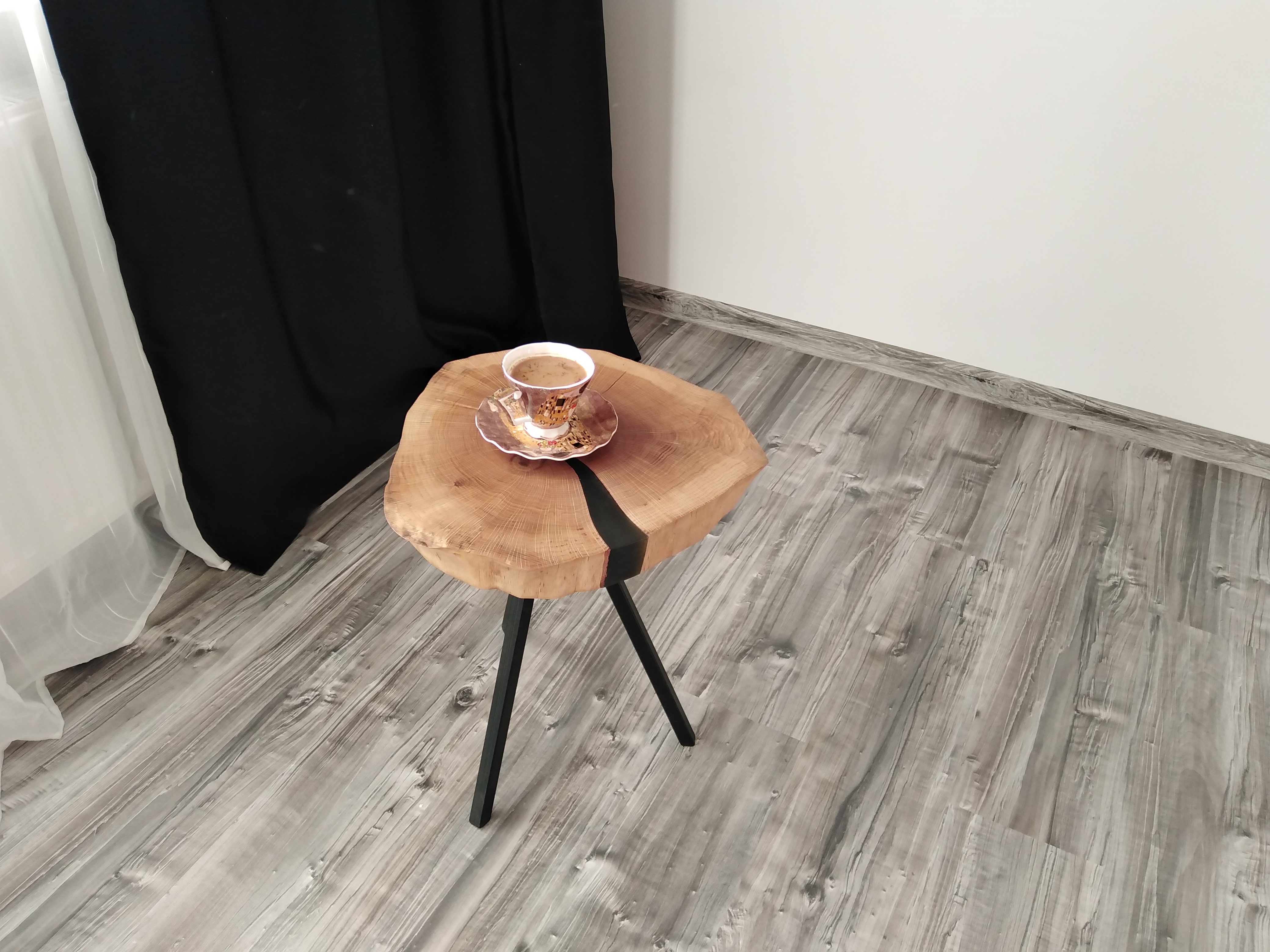 Stolik kawowy z blatem z plastra drewna i czarną żywicą epoksydową w Krakowie, na którym stoi filiżanka kawy, na tle zasłon.