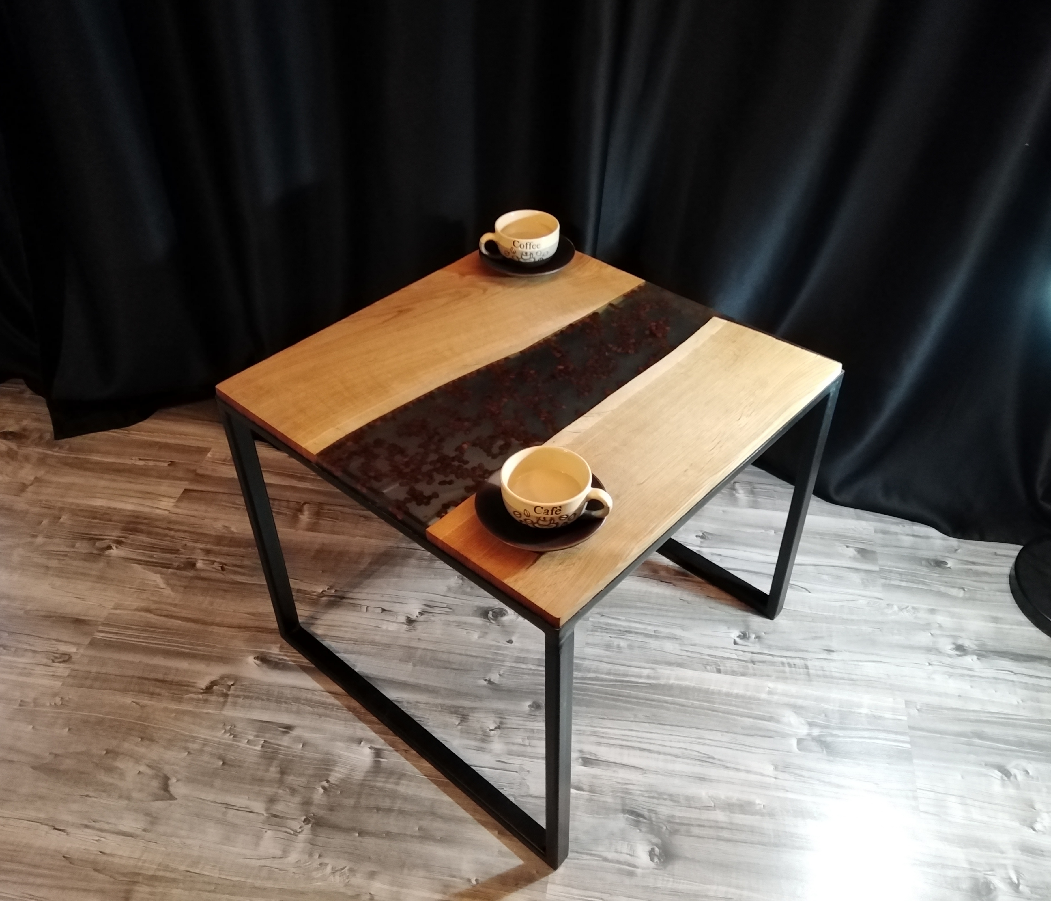 Stolik kawowy z drewnianym blatem i żywicą epoksydową z zatopionymi ziarnami kawy, czarne metalowe nogi, dwie filiżanki kawy na blacie, tło z czarnej zasłony.