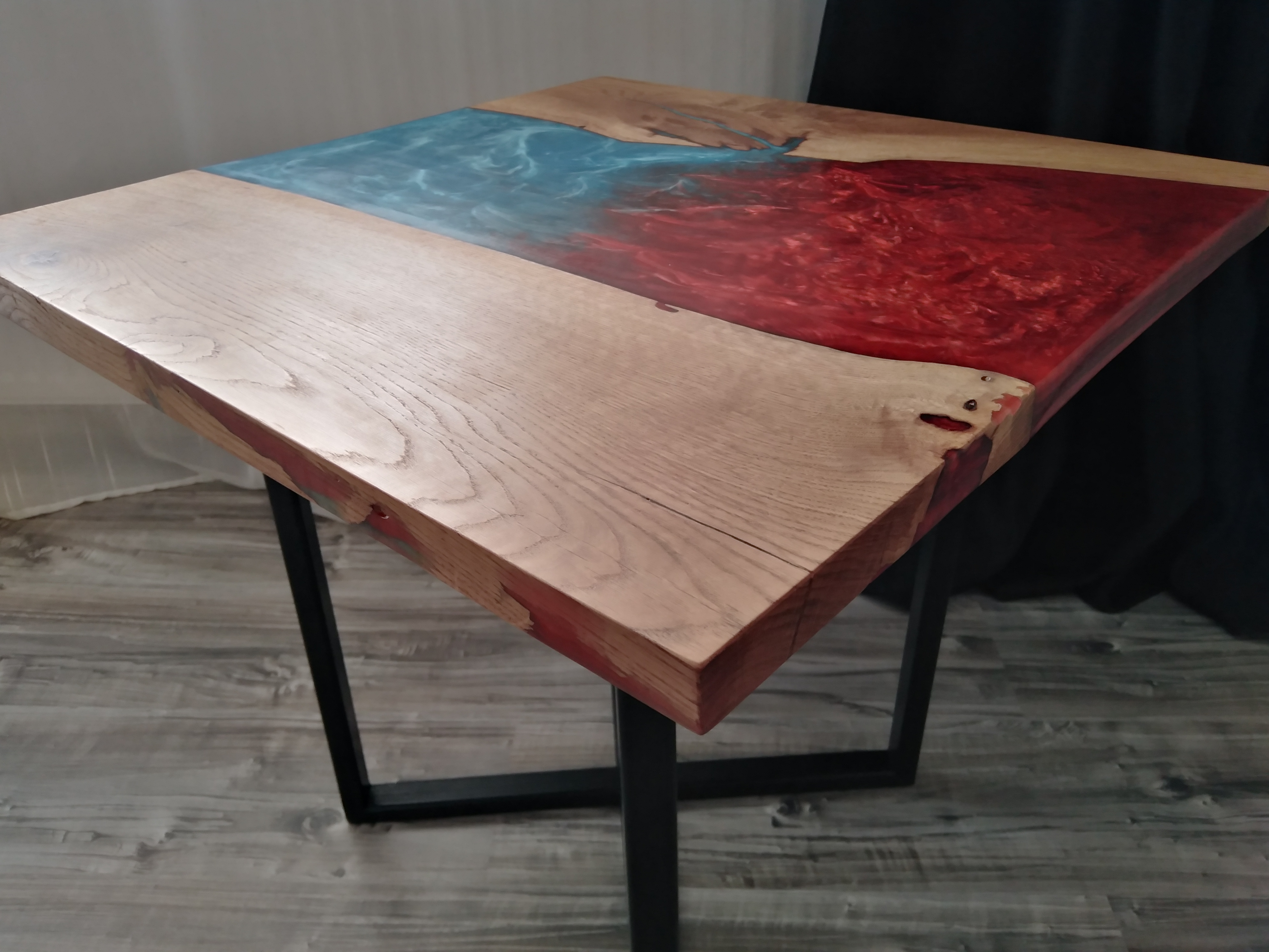 Kwadratowy stolik kawowy z blatem z jasnego drewna z żywicą epoksydową w kolorze czerwonym i niebieskim, osadzony na czarnych, metalowych nogach.